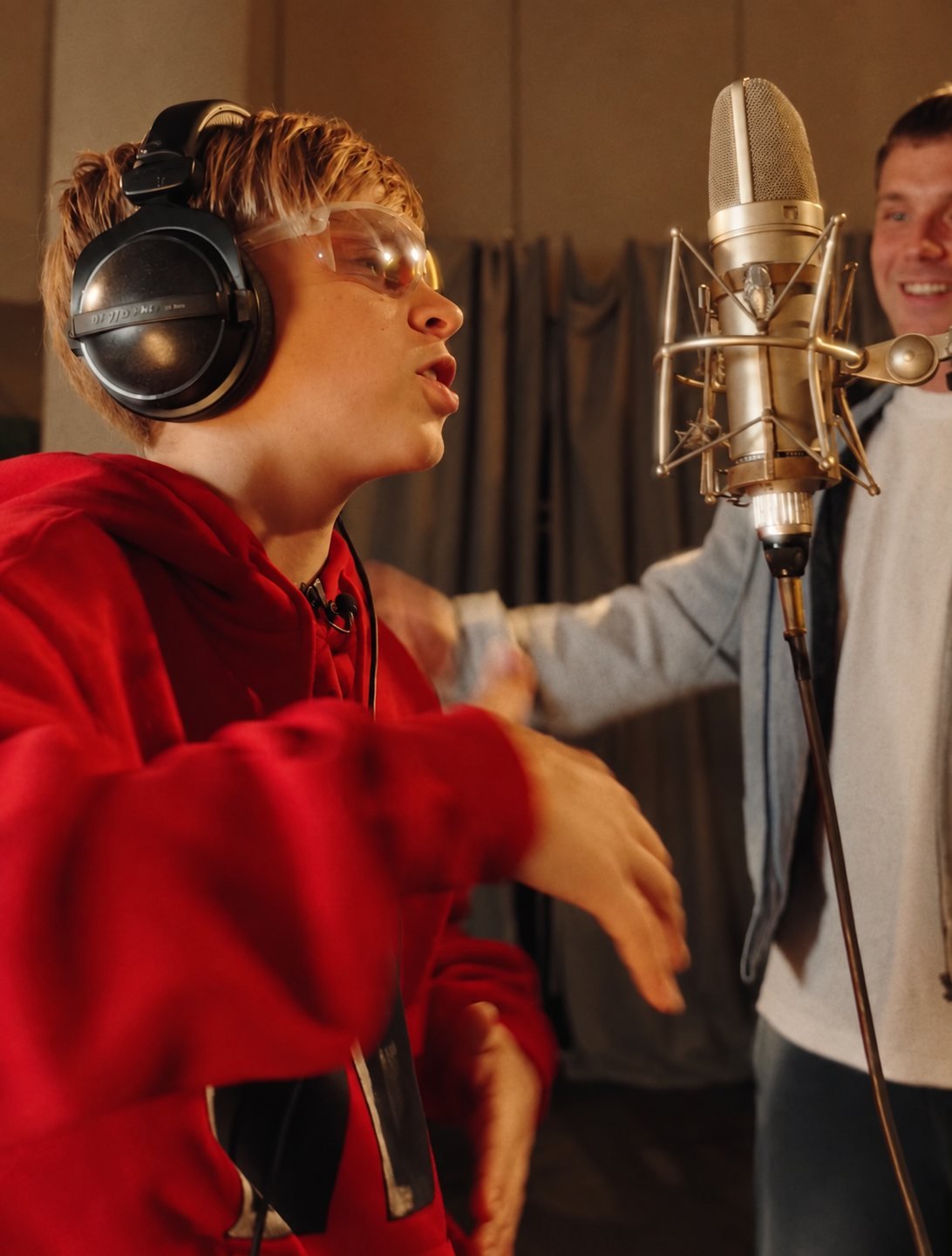 A boy singing into a studio microphone, his mentor smiling beside him